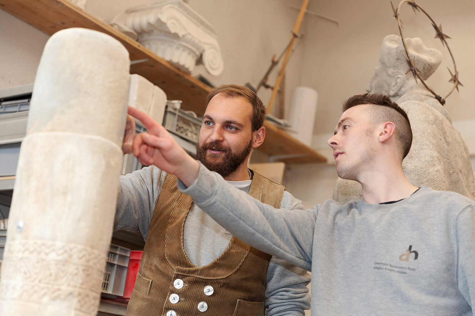 Steinmetz zeigt Lehrling etwas an einer Steinsäule im Atelier