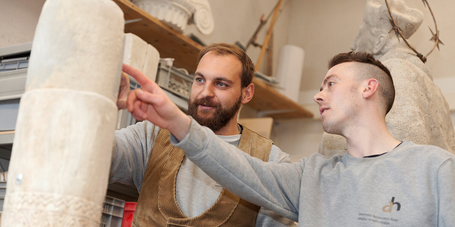 Steinmetz zeigt Lehrling etwas an einer Steinsäule im Atelier