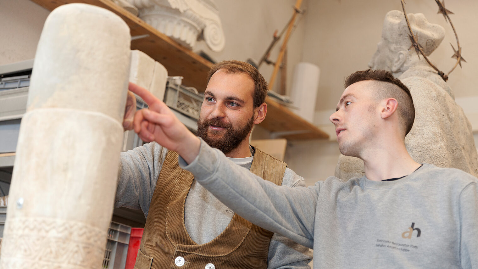 Steinmetz zeigt Lehrling etwas an einer Steinsäule im Atelier