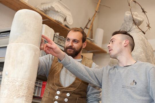 Steinmetz zeigt Lehrling etwas an einer Steinsäule im Atelier