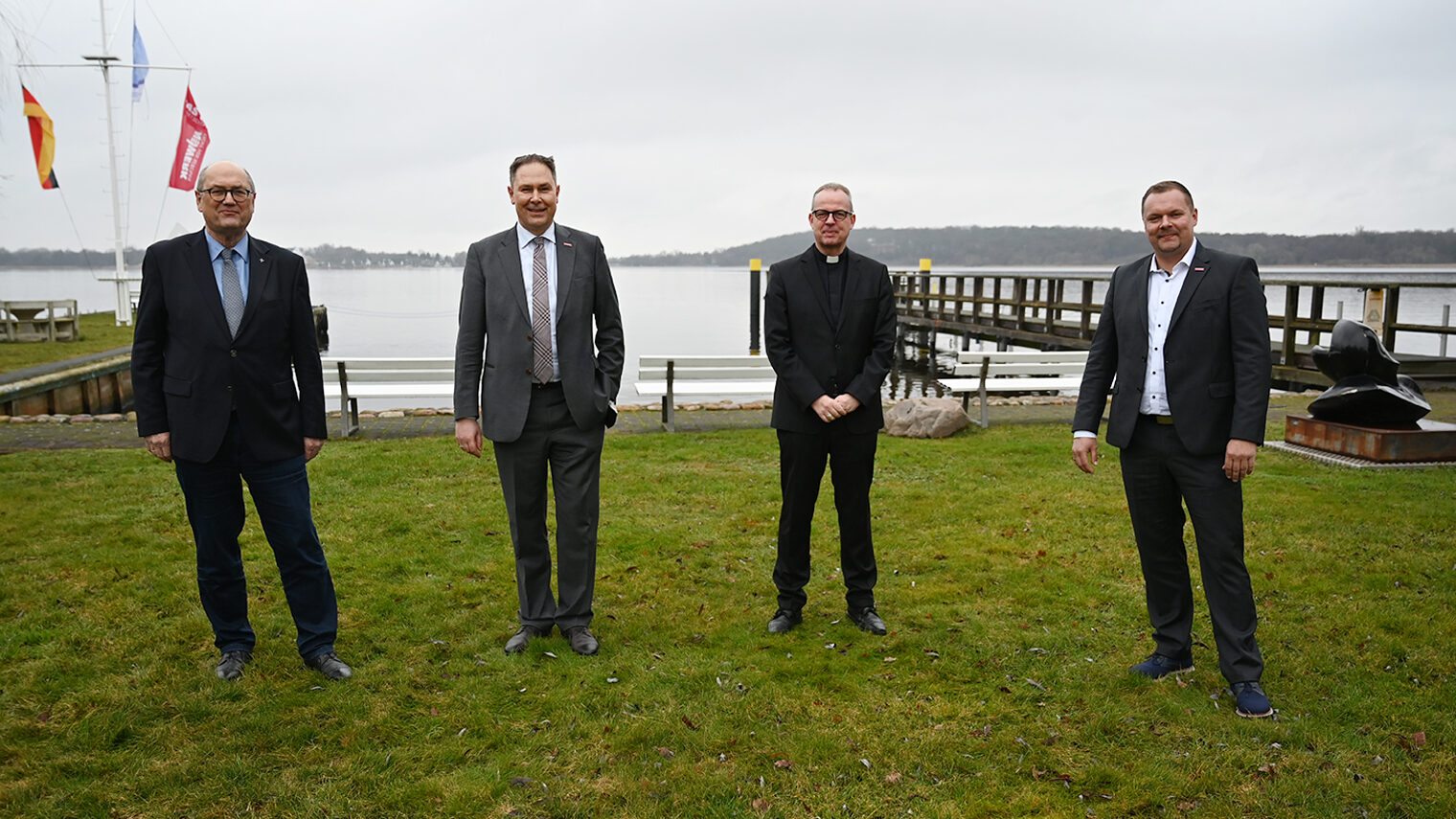 Vier Männer in dunklen Anzügen stehen auf einer Wiese am Seeufer.
