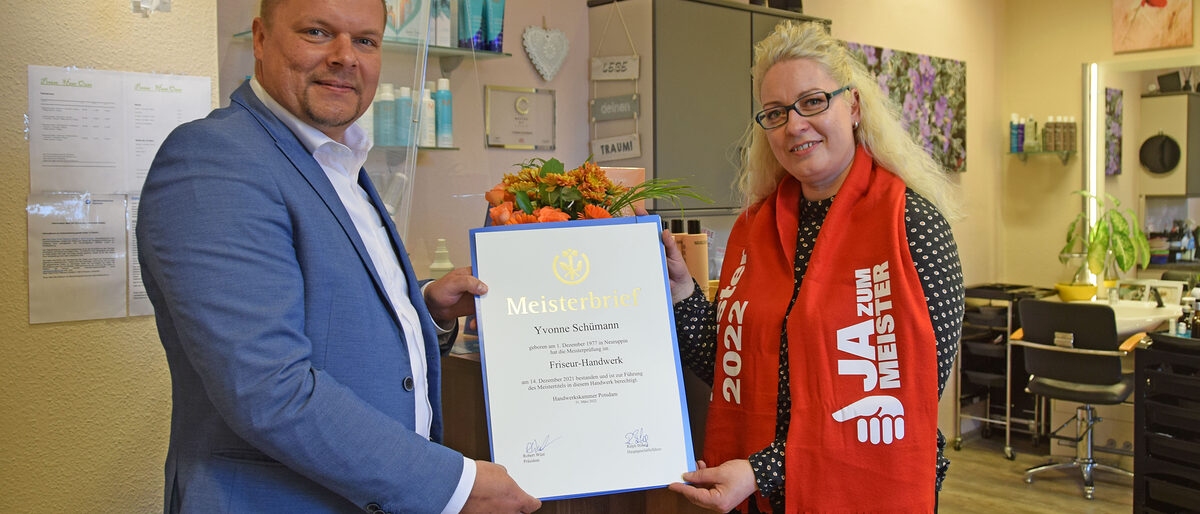 Handwerkskammerpräsident Robert Wüst überreicht Friseurmeisterin Yvonne Schümann den Meisterbrief in ihrem Salon in Wustrau. 2019 ging sie den Weg in die Selbstständigkeit. Mann überreicht junger Frau eine Urkunde im Friseurgeschäft
