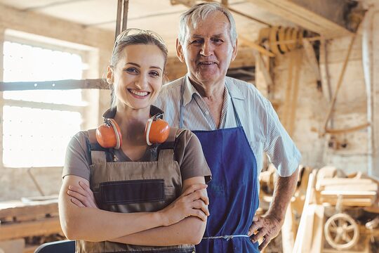 Senior master carpenter with his granddaughter in the wood workshop looking at camera